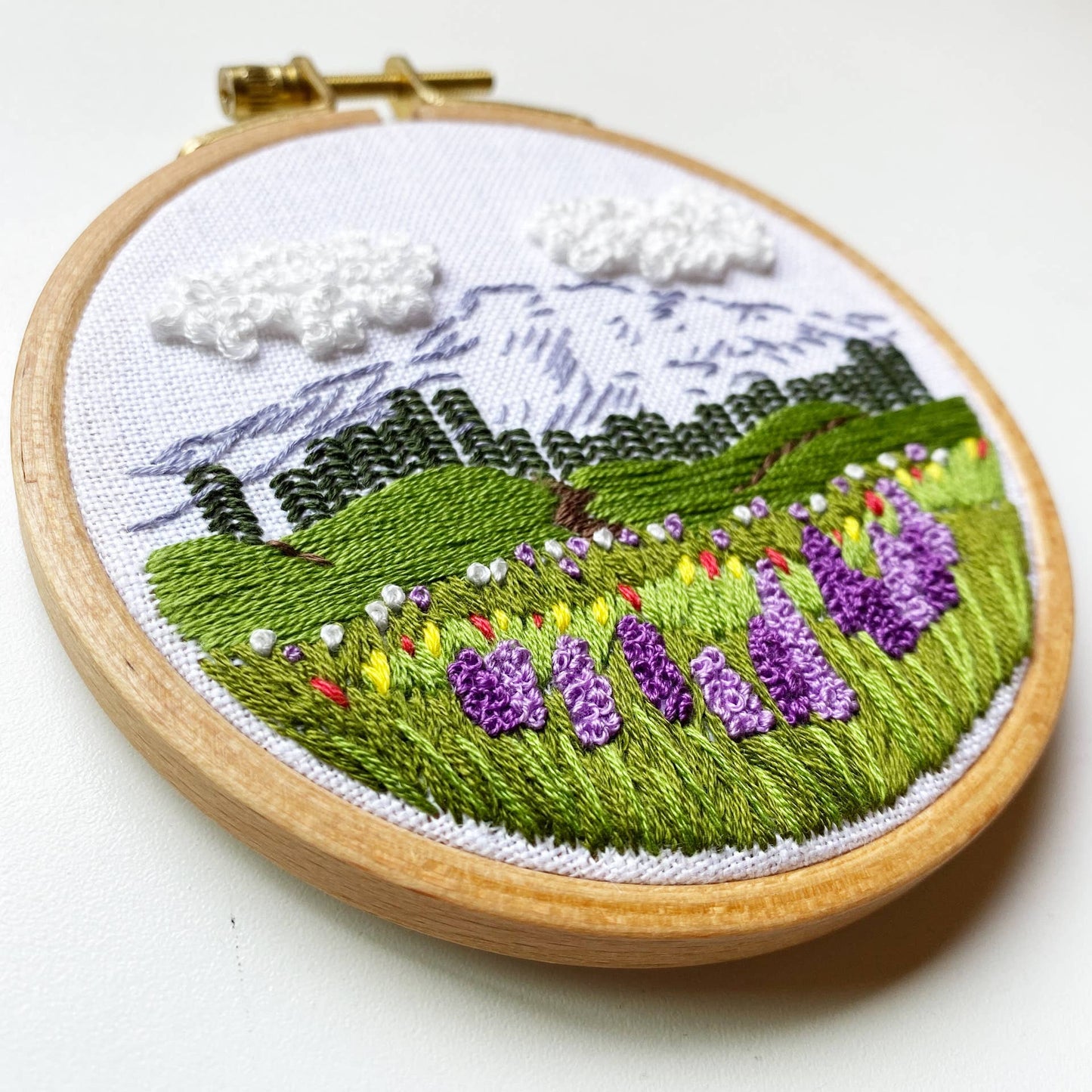 Embroidery hoop with a landscape design featuring green fields, purple flowers, and mountains.