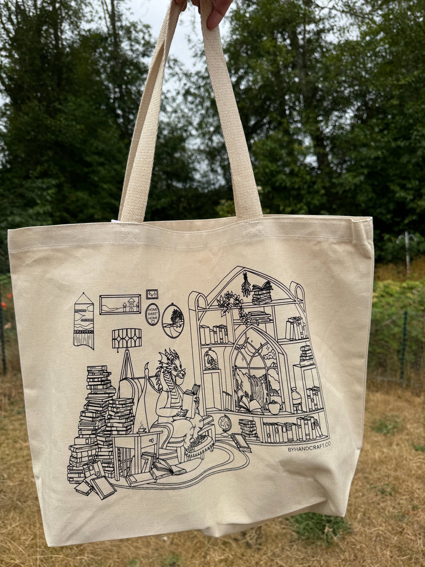 Beige tote bag with black illustration of a dragon in a room filled with books and shelves, held outdoors.