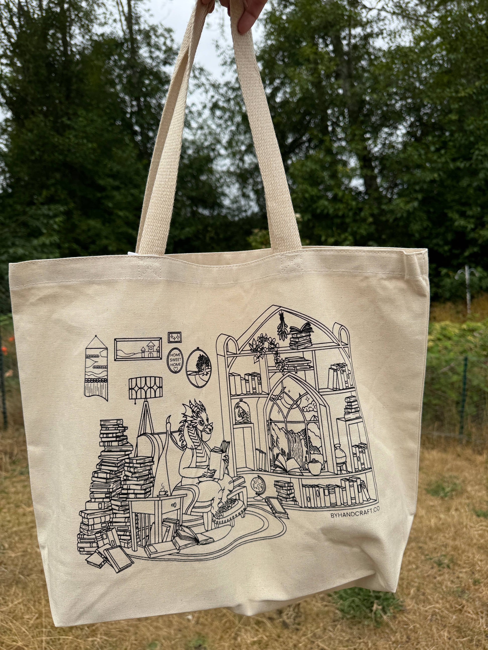 Beige tote bag with black illustration of a dragon in a room filled with books and shelves, held outdoors.