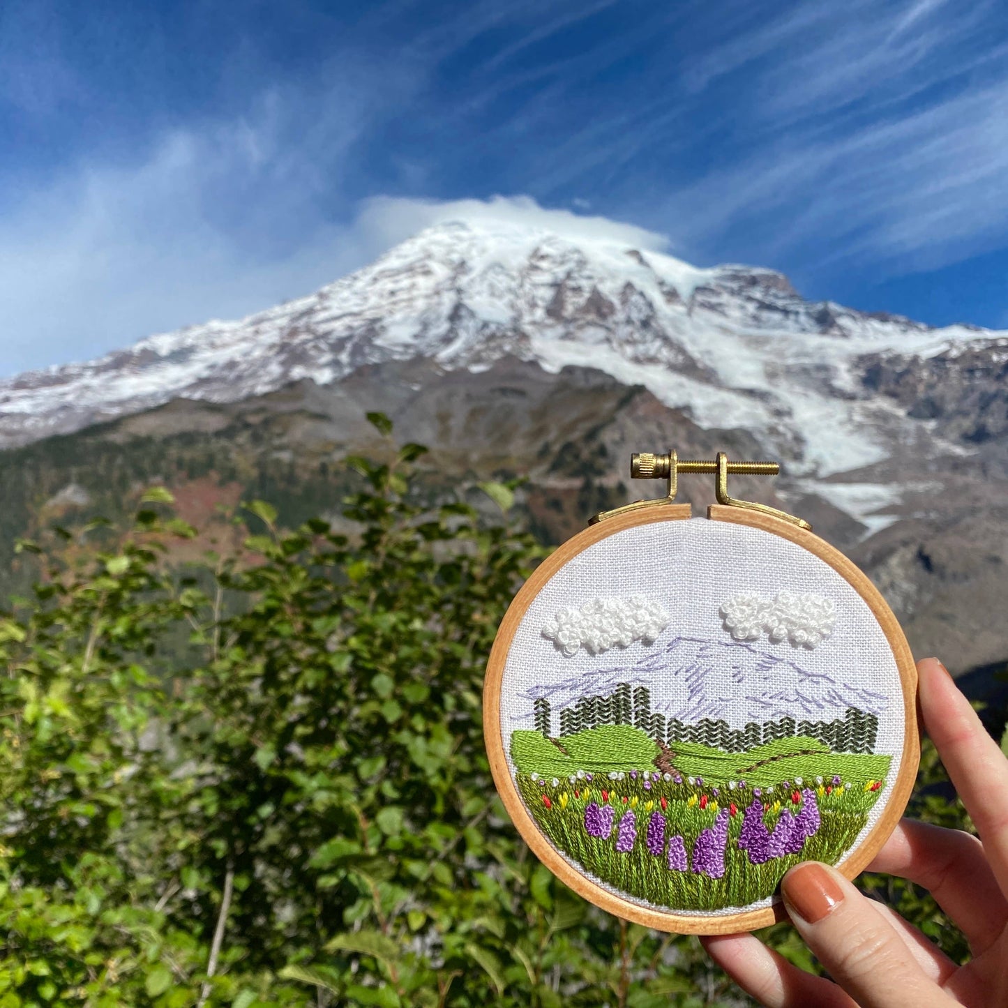 Embroidery hoop with a mountain landscape design held in front of a real mountain.