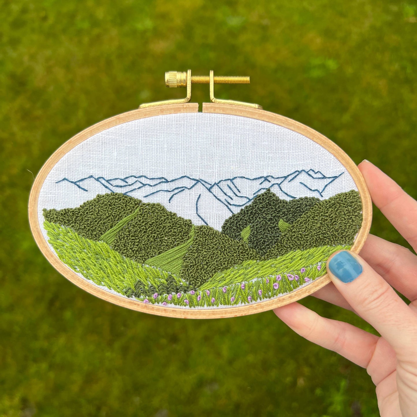 Embroidery hoop with a landscape design held by a hand against a grassy background