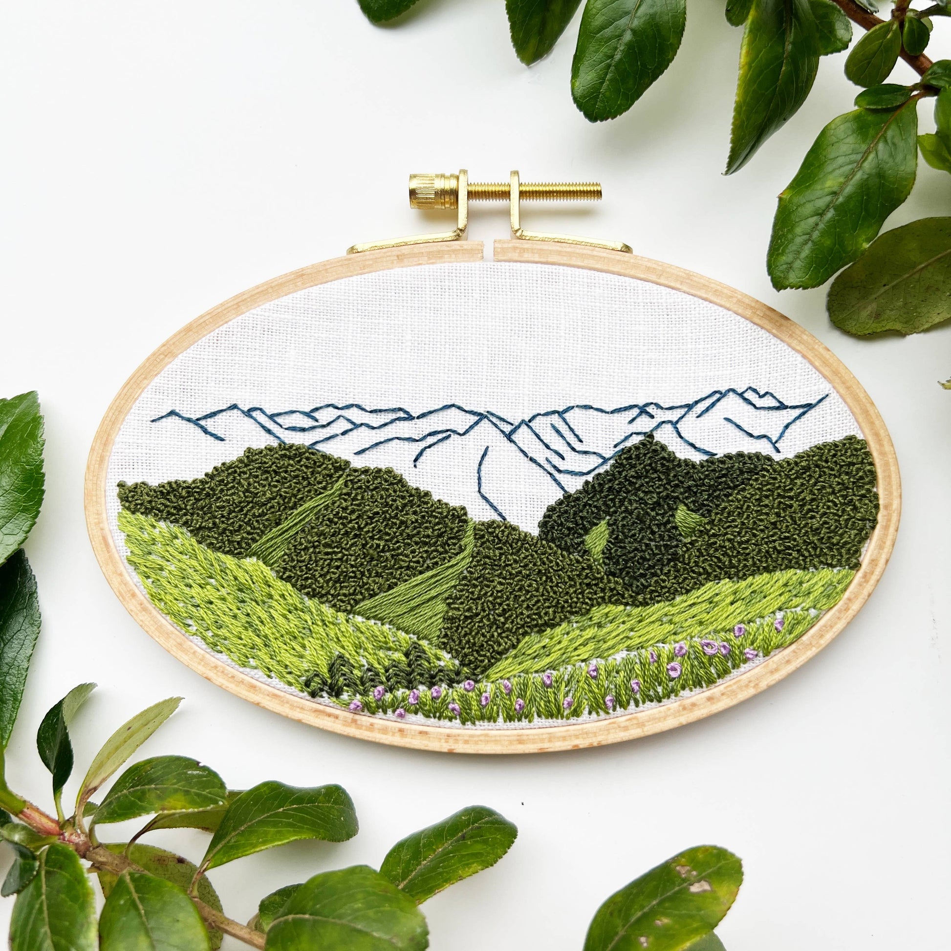 Embroidery hoop with a landscape design featuring mountains and greenery on a white background.