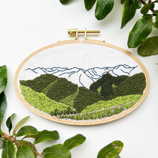 Embroidery hoop with a landscape design featuring mountains and greenery on a white background.