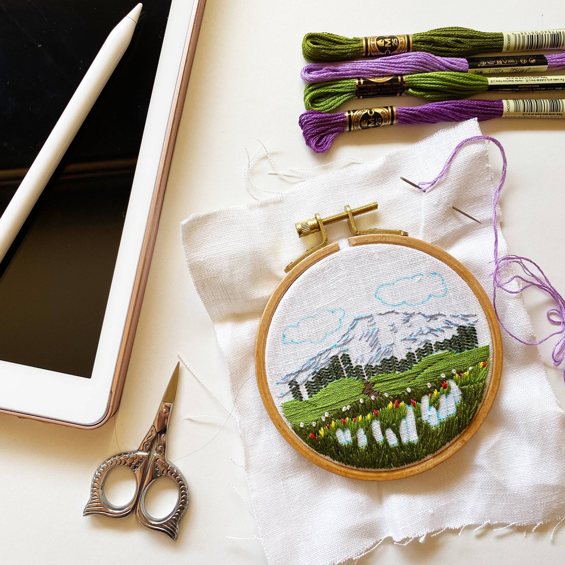 Embroidery hoop with mountain landscape design, embroidery threads, and scissors on a white surface.