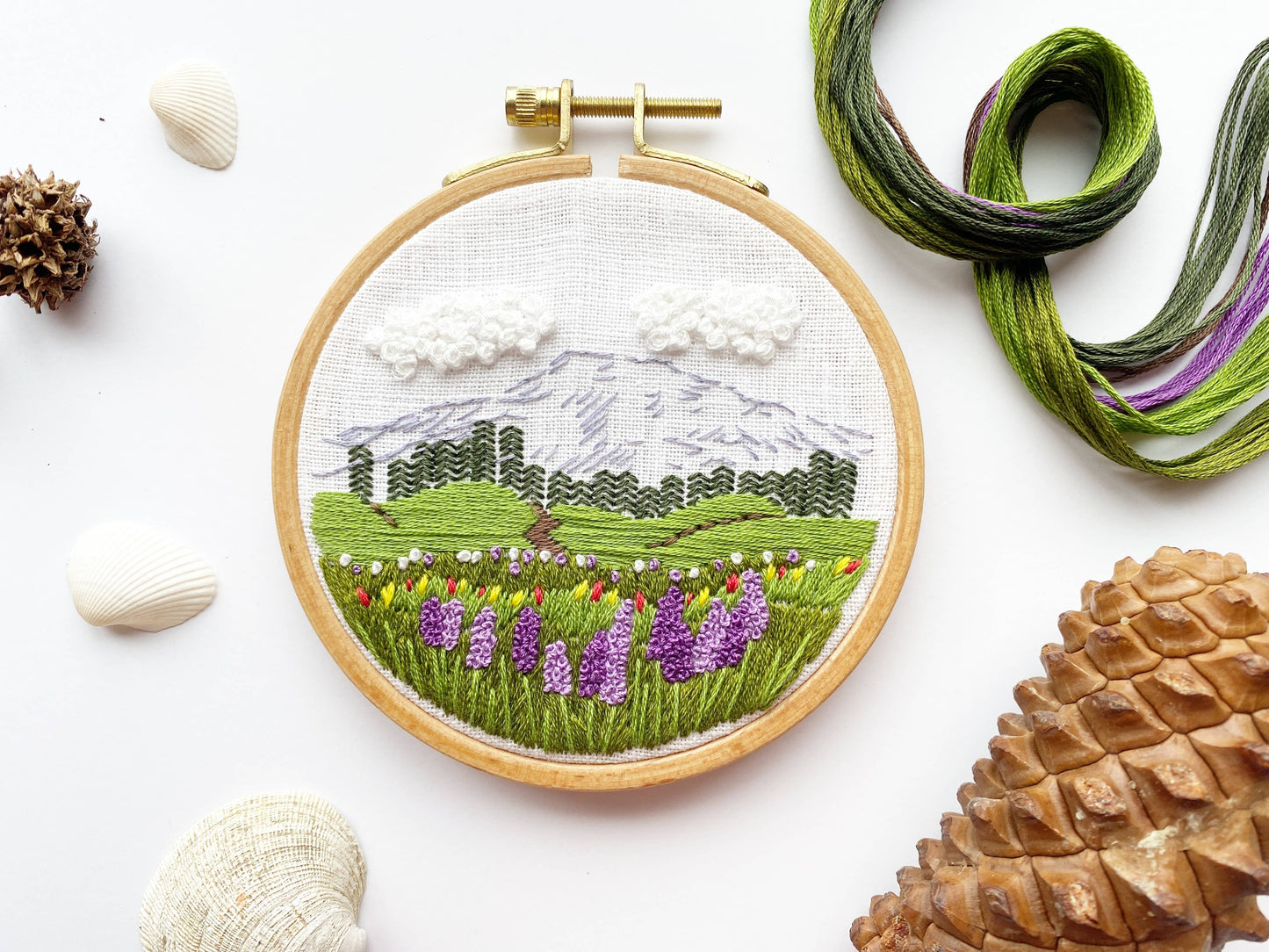 Embroidery hoop with a mountain and landscape design, surrounded by yarn and natural elements on a white background.