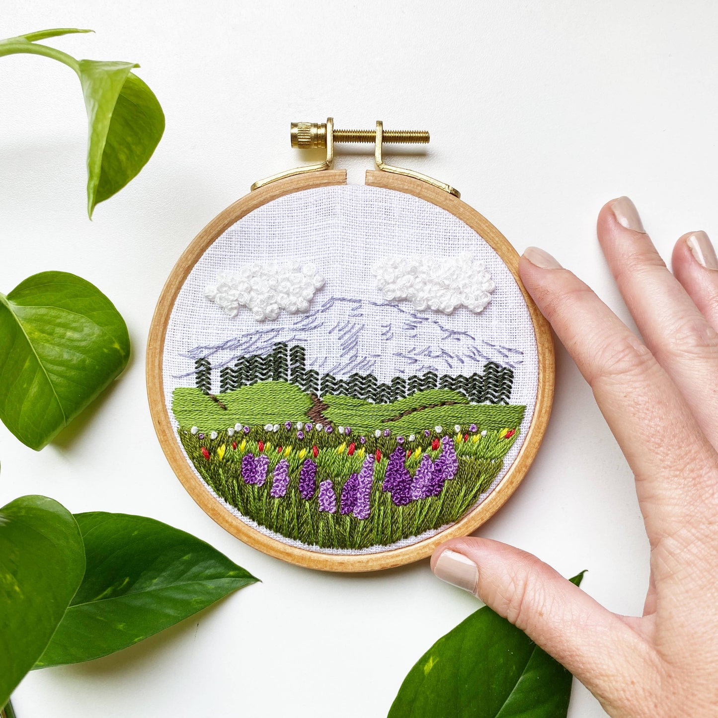Embroidery hoop with a landscape design surrounded by green leaves on a white background
