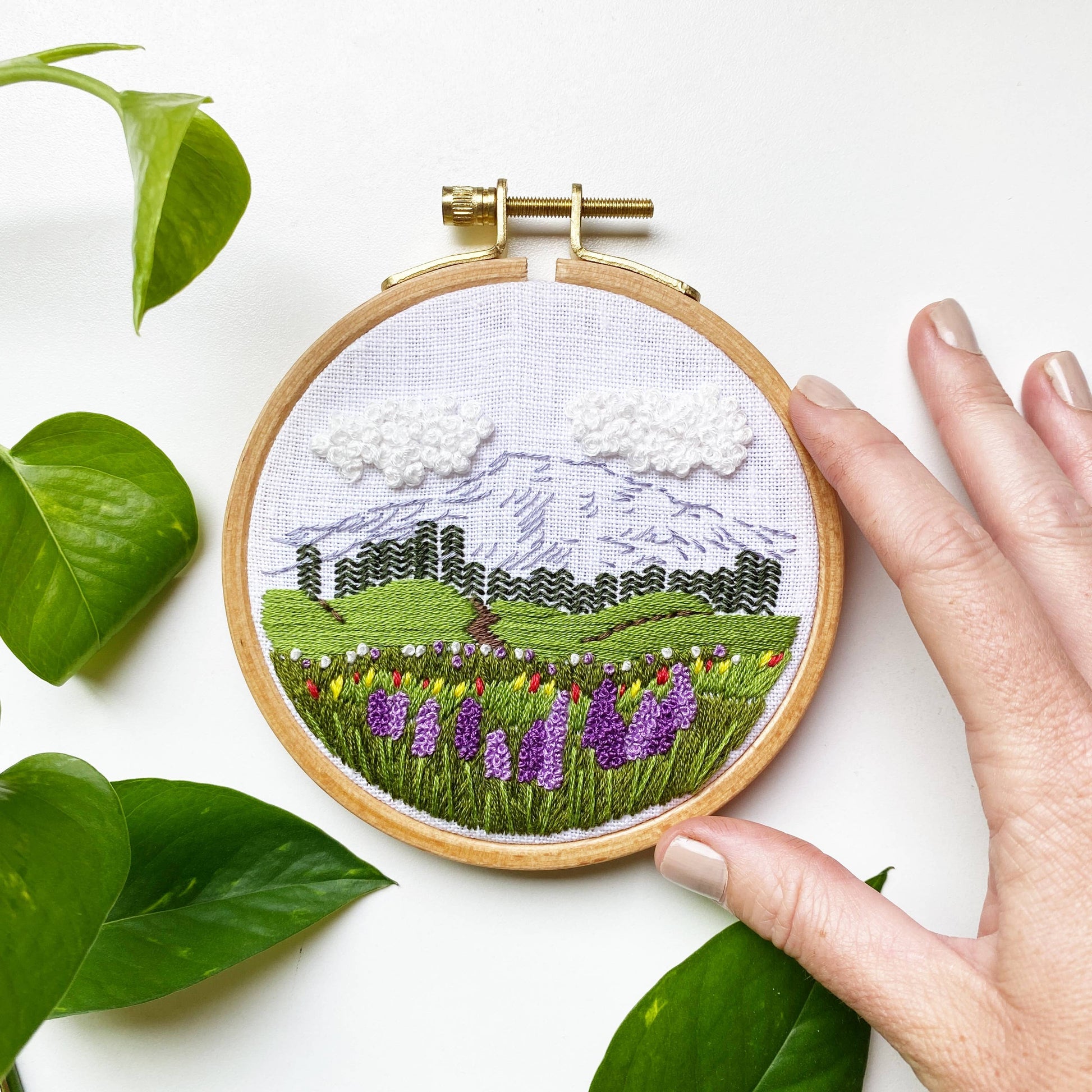 Embroidery hoop with a landscape design surrounded by green leaves on a white background