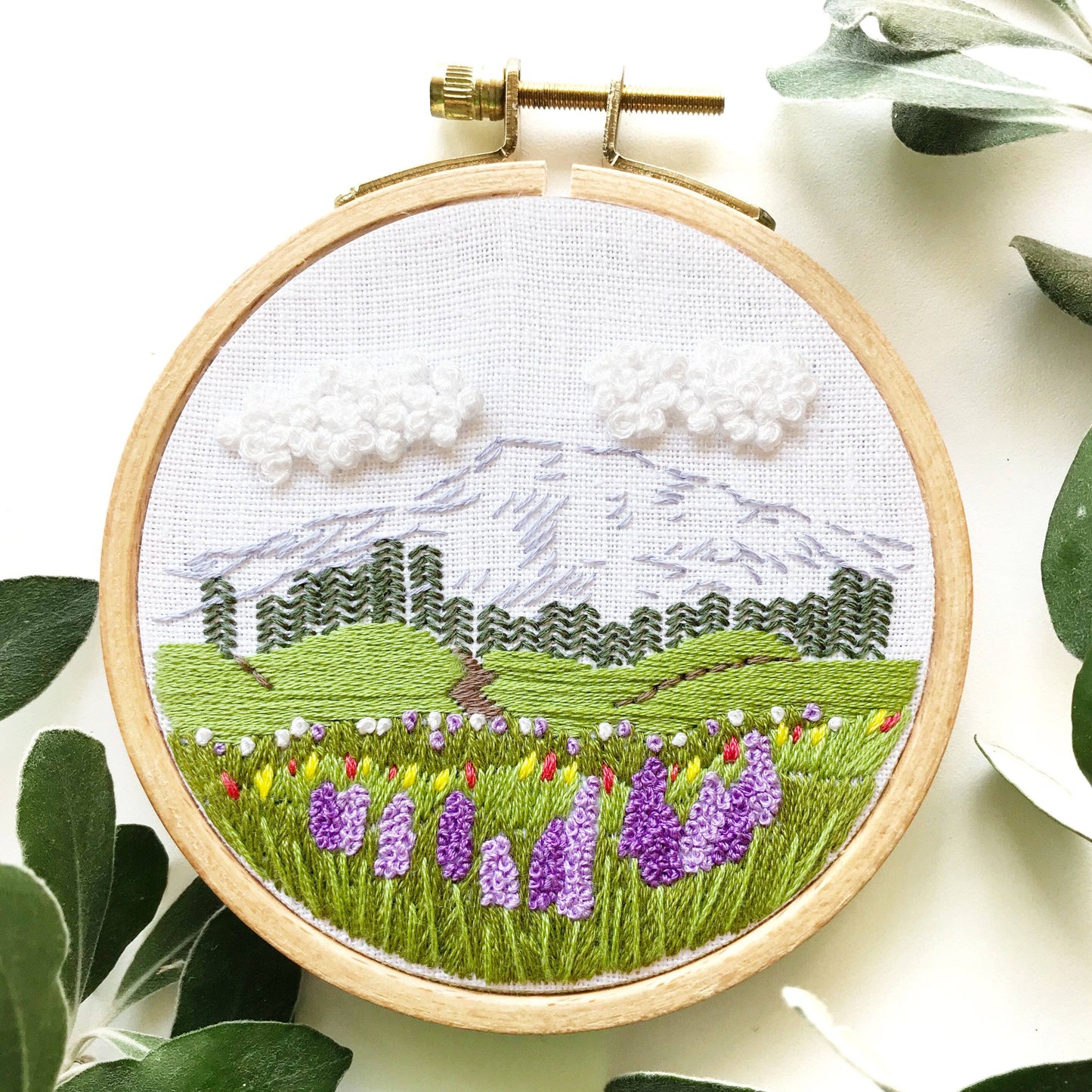 Embroidery hoop with a landscape design featuring mountains, trees, and flowers.