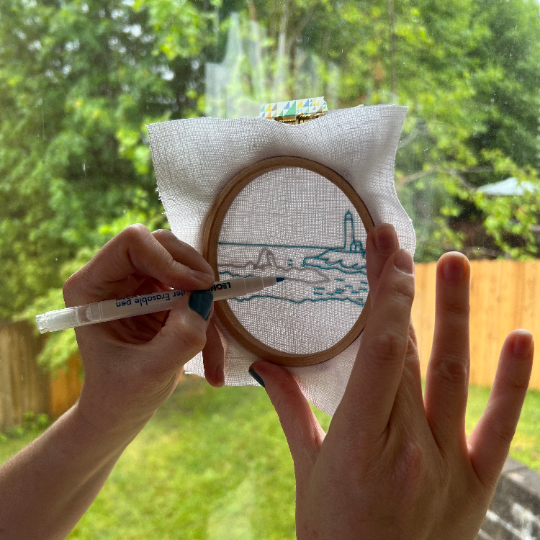 Person holding an embroidery hoop with a partially completed design outdoors.
