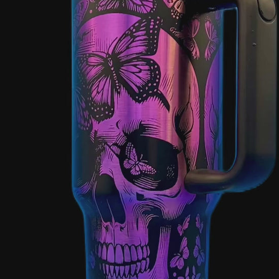 Purple travel mug with black skull and butterfly design, black handle and lid, on a white stand.